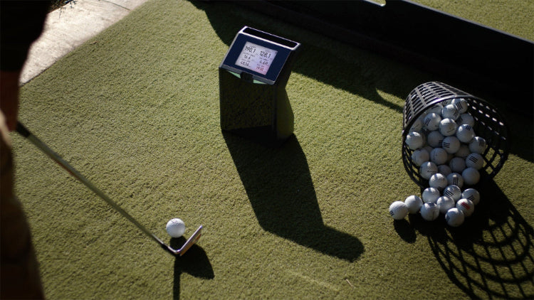 Golf ball on a tee with a digital device and basket of golf balls on a green surface